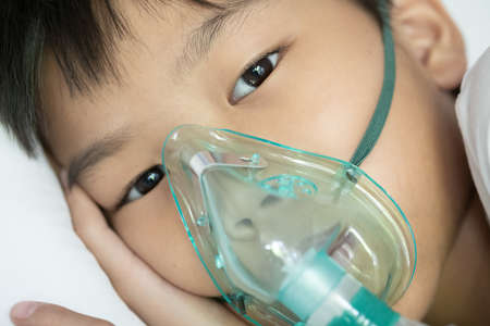 Close up sick Yong asian boy with face oxygen mask lay on a white bed at hospital. COVID 19 concept.の写真素材