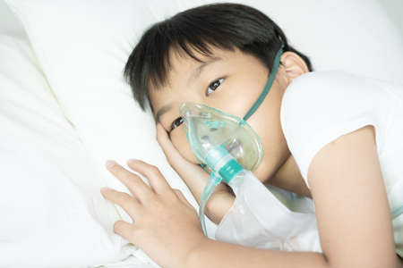 Close up sick Yong asian boy with face oxygen mask lay on a white bed at hospital. COVID 19 concept.の写真素材