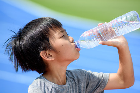 Young Asian boy drink water from a plastic bottle under the sunlight beside a running track to reduce his thirsty.の写真素材