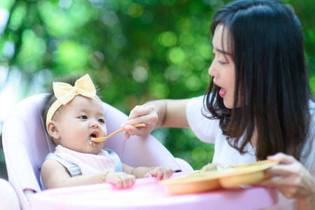 Mother feeding baby infant daughter toddler on highchair in a gardenの写真素材