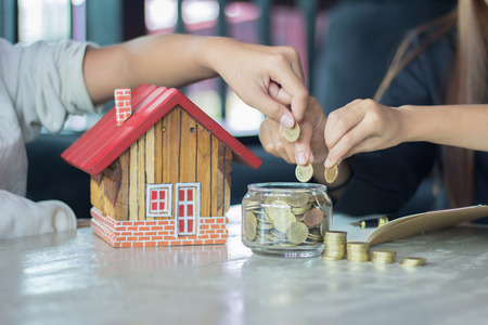 Human hands insert coin in a glass jar, Savings money jar full of coins concept for saving or investment for a house.の写真素材