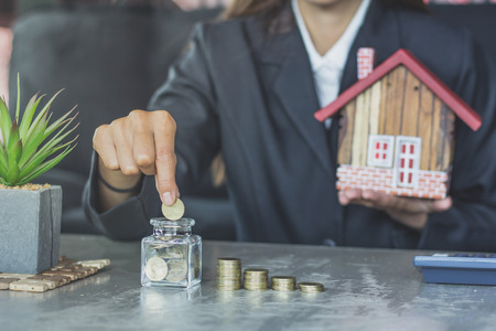 Human hands insert coin in a glass jar and  holding house figure in another , Savings money jar full of coins concept for saving or investment for a house.の写真素材