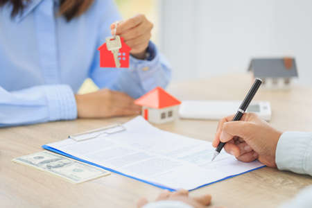 A woman signs a contract to purchase a home with a real estate agent. Model house with keys on table.の写真素材