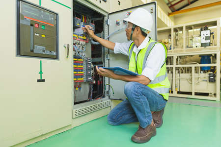 A helmeted technician holds a clipboard while checking and taking notes on the electrical control...