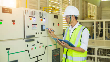A helmeted technician holds a clipboard while checking and taking notes on the electrical control...