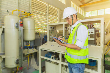 A helmeted technician holds a clipboard while checking and taking notes on the electrical control...