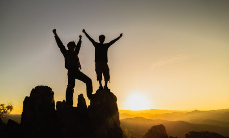 Silhouette of  Hikers climbing up mountain cliff.  Teamwork of two men hiker helping each other on top of mountain climbing team beautiful sunrise landscape.の写真素材
