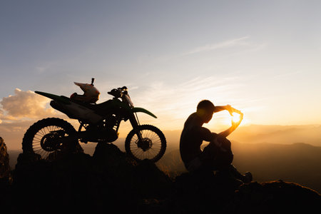 Man with motocross bike against beautiful lights, silhouette of a man with motocross motorcycle On top of rock high mountain at beautiful sunset, enduro motorcycle travel concept.の写真素材