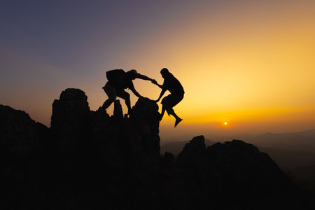 Silhouettes of two people climbing on mountain and helping. Teamwork of two men hiker helping each other on top of mountain climbing team beautiful sunrise. Help and assistance concept.の写真素材