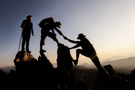Silhouette of  Hikers climbing up mountain cliff. Climbing group helping each other while climbing up in sunset. Concept of help and teamwork.の写真素材