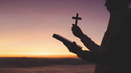 Silhouette of woman praying while holding Bible and cross, Pray in the Morning , Woman praying with hands together on the Sunrise background.の写真素材