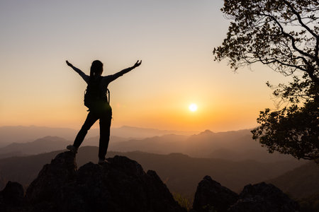 Silhouette of a person standing on top of the mountain with their arms raised in victory, overlooking misty mountains and clouds at sunrise.の写真素材
