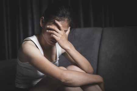 A depressed young woman sits sadly on a sofa, covering her face with her hands. Depressing atmosphere. Relationship problems. Violence against women.の写真素材