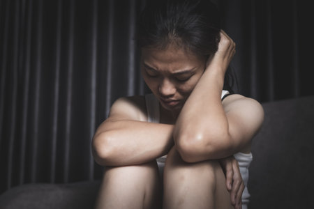 A depressed young woman sits sadly on a sofa, covering her face with her hands. Depressing atmosphere. Relationship problems. Violence against women.の写真素材