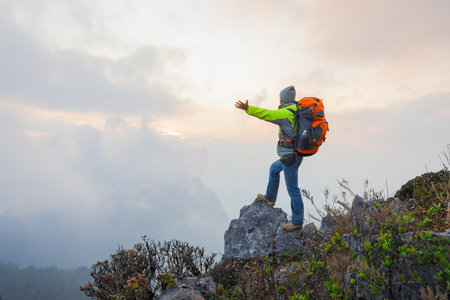 A lone man hiker with backpack  stands on a rocky peak, gazing out at a breathtaking vista of mountains bathed in the warm glow of the setting sun,  Concept motivation and goal achievement.の写真素材