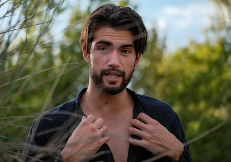 portrait of beautiful dark-haired italian model with light eyes and black shirt with beard in the trees. High quality photoの写真素材