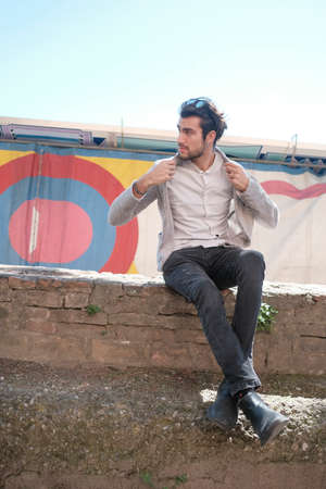 handsome Italian dark-haired guy with jacket sitting on low wall in sunny day. High quality photo