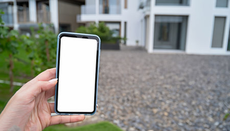 Mockup image of a hand holding mobile smart phone with white screen in front of houseの素材