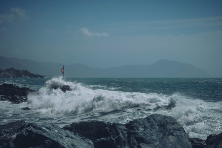 Powerful waves crash against dark rocks in a rough sea, with a distant horizon under a cloudy sky.