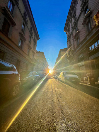 Straight-on view down a narrow Milan street with buildings on either side. Sunlight bursts through the street, creating a lens flare and casting long shadows. High quality photoの写真素材
