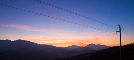 A serene landscape featuring silhouetted mountains against a twilight sky with hues of orange and blue, punctuated by power lines. High quality photoの写真素材