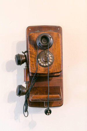 An antique, wooden, wall-mounted telephone with a rotary dial and separate earpiece and mouthpiece, displayed on a light background. High quality photoの写真素材