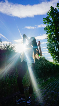 A woman stands with her arms raised, silhouetted against the bright sun and a clear blue sky,...