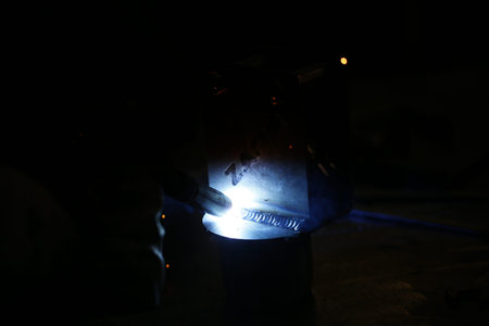 A bright, intense welding arc illuminates a dark workshop as someone welds metal, with sparks flying...
