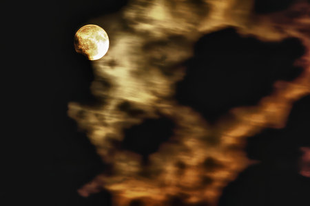A vibrant, glowing full moon is partially veiled by dark and illuminated golden clouds, creating a...