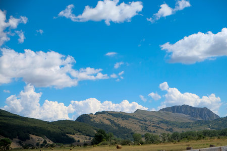 A wide shot of a vast field with numerous round hay bales scattered across the ground, all set against a large, green, sunlit mountain. in Abruzzo, Italy. High quality photoの写真素材