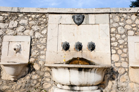 An ancient stone fountain featuring a large basin and three carved grotesque faces, set against a stone wall with a coat of arms. High quality photoの写真素材
