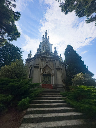A striking, low-angle view of an ornate Gothic mausoleum with a grand stone staircase, surrounded by lush green foliage under a dramatic blue sky. High quality photoの写真素材