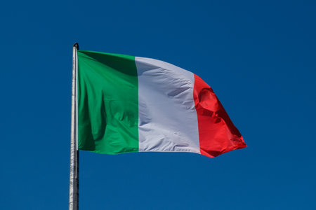 The Italian tricolor flag, with its green, white, and red stripes, waves gracefully in the wind against a clear and bright blue sky. High quality photoの写真素材