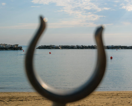 A calm seascape viewed through the out-of-focus metal oarlock or rowlock of a beach chair boat, focusing on the sandy beach, blue water, and horizon with breakwaters. High quality photoの写真素材