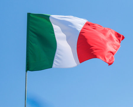 The Italian tricolor flag green, white, and red waving proudly on a flagpole, set against a smooth, bright and clear blue sky background. Symbolic of national pride and identity. High quality photoの写真素材