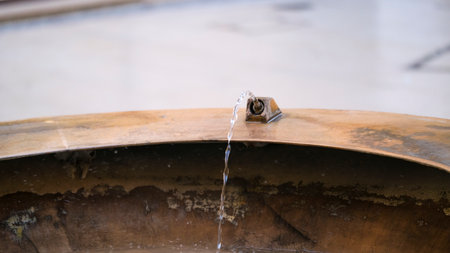 A serene close-up shot of a small stream of water falling from an aged metal nozzle into a large, curved basin. The bronze-toned surface shows visible patina and wear. High quality photoの写真素材