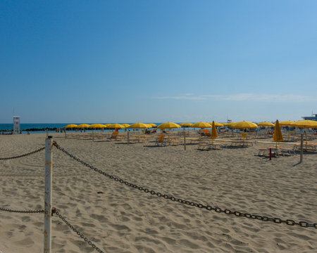 A wide shot of a neatly organized, empty sandy beach under a clear blue sky, featuring rows of bright yellow umbrellas and matching chaise lounges ready for summer tourists. High quality photoの写真素材