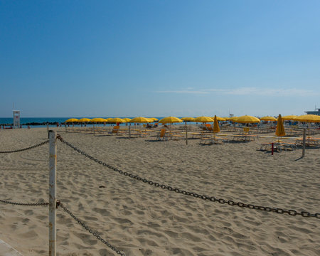 A wide shot of a neatly organized, empty sandy beach under a clear blue sky, featuring rows of bright yellow umbrellas and matching chaise lounges ready for summer tourists. High quality photoの写真素材