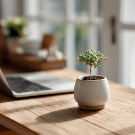 A close-up, bright, and minimalist photograph of a small potted plant on a light wooden desk, with a ceramic mug and laptop in the soft-focus background, suggesting a clean workspace. High quality photoの素材