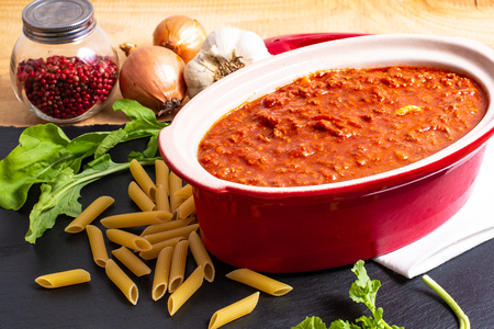Homemade slow cooker bolognese sauce in red ceramic pot and raw penne, peppercorn, garlic, onion and rocket saladの写真素材