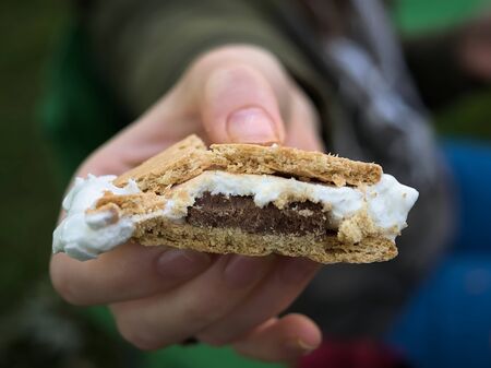 A woman/man hands a perfectly cooked Smore to the viewer. Smore in focus smashed smooshed showing the gooey marshmallow chocolate crumbling crackerの写真素材