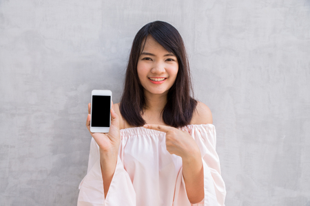 Smiling woman showing a blank smartphone screen standing on concrete wall.の写真素材
