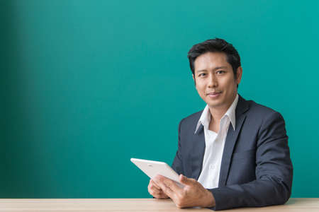 Portrait of businessman smiling and using pad at desktop against of blue wall. Vintage filtered image.の写真素材