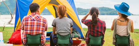 Group of man and woman enjoy camping picnic and barbecue at lake with tents in background. Young mixed race Asian woman and man. Panoramic banner.の写真素材