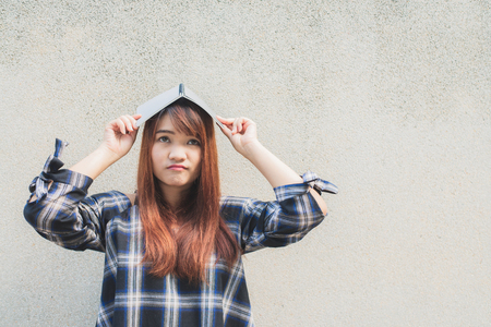 Smiling young beautiful asian woman thinking with a book over head on concrete wall background. Vintage effect style pictures.の写真素材