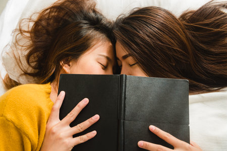 Top view of beautiful young asian women lesbian happy couple kiss and smiling while lying together in bed under book at home. Funny women after wake up. Lesbian couple together indoors concept.の写真素材