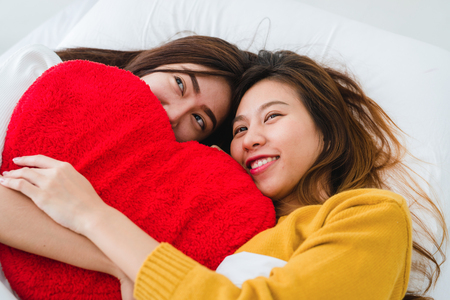 Top view of beautiful young asian women lesbian happy couple hugging and smiling while lying together in bed under blanket at home. Funny women after wake up. Lesbian couple together indoors concept.の写真素材