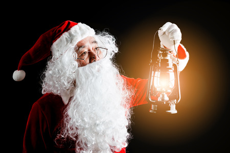 Photo of happy Santa Claus with a lantern of Christmas present in hands, isolated on black background.の写真素材