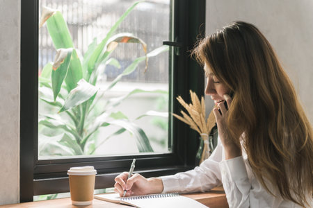 Young business woman in white dress sitting at table in cafe and writing in notebook. Asian woman talking smartphone and cup of coffee. Freelancer working in coffee shop. Student learning online.の写真素材