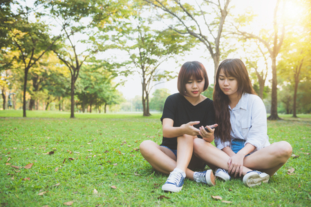Two beautiful happy young asian women friends having fun together at park and taking a selfie. Happy hipster young asian girls smiling and looking at smartphone. Lifestyle and friendship concepts.の写真素材